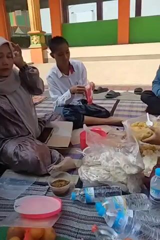 This is a video of a gathering of people, likely outdoors, with a building visible in the background. The people are seated on the ground on mats, with food and drinks scattered around them. Several individuals are wearing hijabs, and some men are wearing skullcaps. One woman is adjusting her hijab, and a young boy is holding a bottle of water. In the background, more people are seated, and some are standing and walking. The building appears to be a school or a place of worship, with orange and green accents on its exterior. The overall impression is of a casual, communal gathering.