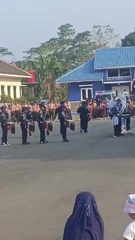 The video shows a marching band performance outdoors in front of a blue building. A group of young people, dressed in dark uniforms with red accents, are playing drums. They are arranged in a line and appear to be in formation. Behind them, a larger group of people, many in brown uniforms, are standing and watching. Some individuals in the foreground are also observing the performance. The environment includes pavement, some trees, and a flagpole. The sky is visible in the background, suggesting it is daytime. The overall scene depicts a school or community event.