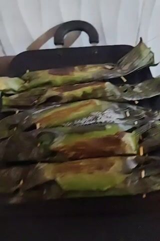 The video shows a black griddle pan containing six green-and-brown packages. The packages are arranged in two rows of three, and they are held together by wooden skewers. The pan has a black handle. A white curtain is visible in the background.