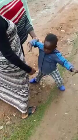 The video shows a young child, possibly a toddler, standing in a muddy area by the side of a road. The child is wearing a blue jacket, plaid pants, and blue shoes. Two women are standing near the child, with one holding the child's arm. The women appear to be helping the child navigate the muddy ground. The woman holding the child's arm is wearing a shawl-like garment with red and white stripes, while the other woman is wearing a black and white patterned dress. The background shows a dirt road and some vegetation. The child appears to be holding something in his hand.