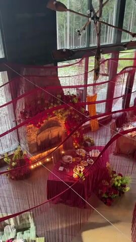 This is a video showing an interior decorated for an event, likely a celebration or intimate gathering. The scene is set in a room with large windows overlooking a wooded area, suggesting a location in Kanabekovo, Russia. The time of day appears to be evening or late afternoon, as the lighting is dim and accent lights are used to create a warm ambiance. The weather outside is not discernible.

The room is draped with flowing red fabric, creating a dramatic and festive atmosphere. The focal point of the room is a large stone fireplace, adorned with abundant red and yellow floral arrangements. In front of the fireplace, a small, round table is set with a red tablecloth. It is laden with food items, including pastries, fruits, and what appear to be small plates of appetizers. Candles are also placed on the table, casting a soft glow.

Around the table, there are additional floral arrangements in complementary colors of red, green, and yellow, adding to the decorative theme. In the background, other tables are partially visible, also draped in fabric and suggesting further setup for guests. A person can be seen walking in the background through the large windows, indicating that the event may be ongoing or in its final preparations.

The overall mood conveyed by the video is one of warmth, celebration, and perhaps a touch of romance, due to the rich colors, soft lighting, and abundant flowers. There is no visible text in the media.