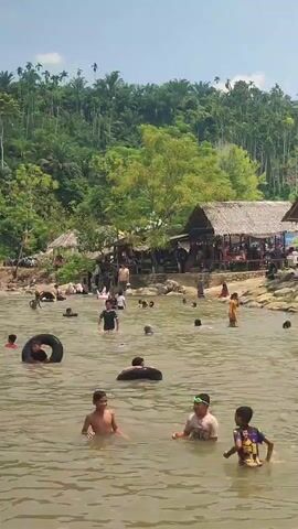 This is a video taken from a slightly elevated perspective looking down at a shallow, muddy river. Numerous people, primarily children and some adults, are enjoying themselves in the water. Several individuals are using inflatable tubes, while others are swimming or wading. In the background, along the riverbank, are several thatched-roof huts or shelters, under which groups of people are gathered. Beyond the shelters, a lush green hillside covered in dense vegetation and trees rises steeply. The sky is clear and bright, indicating a sunny day. The overall scene depicts a lively and crowded recreational area, likely a popular spot for swimming and socializing.