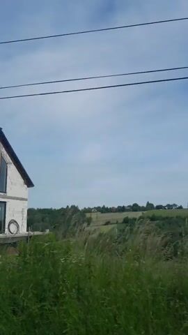 The video shows a series of exterior shots of houses under construction in a rural area. The first shot shows a modern house with large windows and a dark roof. The second shot shows a similar house with a wooden door and some blue tarps. The third shot pans to reveal more houses and a large evergreen tree. The last shot focuses on the evergreen tree. The sky is cloudy and there is some construction debris visible in the foreground.