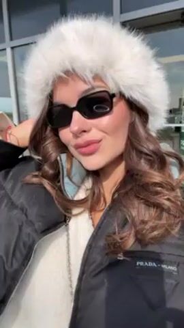 A woman with long, curly brown hair is standing outside, wearing a white fur hat, black sunglasses, a white sweater, and a black Prada puffer jacket. She blows a kiss to the camera with her right hand, which has a ring on the ring finger. She is in front of a glass-walled building with a yellow sign on the door. The sun is shining brightly.