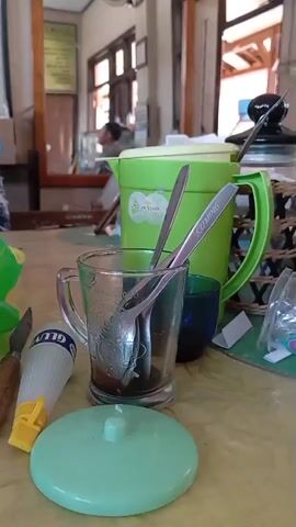 This is a close-up, slightly blurry, eye-level shot of a table in what appears to be an indoor eating establishment. In the foreground, on the table, is a clear glass with a handle containing two spoons. To the left of the glass is a tube of glue with a yellow cap and a silver tip, and a light green lid. Behind the glass is a tall, bright green pitcher with a yellow lid and a dark green handle. Behind the pitcher, to the right, is a woven basket containing several glass jars with dark lids, and what looks like a plastic bag. In the background, out of focus, is a person sitting at a counter and several wooden doors and windows. The overall impression is of a casual, possibly outdoor or semi-outdoor, dining area.