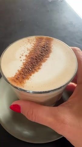 A close-up shot focuses on a person's hand with bright red nail polish holding a clear glass cup filled with a frothy latte. The latte is topped with a dusting of cinnamon or cocoa powder, artfully arranged in a diagonal stripe across the foam. The cup rests on a light grey saucer. The background is dark and textured, suggesting a tabletop, possibly in a cafe or restaurant. In the upper right corner of the frame, a small, white square with a black number "03" is visible, indicating a table number or identifier, further supporting a cafe setting.

The lighting suggests it could be daytime, with a soft light illuminating the drink. The overall mood conveyed is one of simple indulgence and enjoyment of a warm beverage. The hand holding the cup appears relaxed and casual. The Bellingen, Australia location context implies a relaxed, possibly coastal or rural Australian cafe atmosphere.
