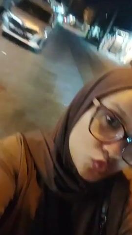 A woman wearing glasses and a hijab is filmed from the chest up as she sits on the back of a motorcycle. She is wearing a brown jacket. The motorcycle is moving at night on a street. In the background, there are lights from buildings and streetlights, and other vehicles on the road.
