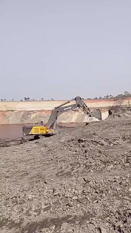 The video shows a large yellow and gray Volvo EC950E excavator digging into a pile of dark gray material that appears to be rock and dirt. The excavator is situated on top of the same material, close to a body of murky water. In the background, there's a large, light-colored embankment with some sparse vegetation visible on top. The sky is light blue. The excavator is actively digging into the pile, scooping up material with its bucket.