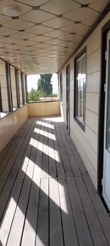 The video shows a view from a covered porch or balcony. The floor is made of wooden planks. The walls are light-colored, and there are windows along one side. The ceiling is tiled with a geometric pattern and has a light fixture. Sunlight casts striped shadows on the floor. Beyond the porch, there are trees and buildings visible under a clear sky. The camera pans slightly to show different parts of the porch and the outdoor view.