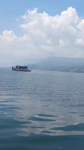 A ferry boat travels across a body of water under a partly cloudy blue sky. The boat is white with a red stripe and has multiple decks. In the background, hazy mountains and hills are visible. The water in the foreground has ripples reflecting the sky and clouds.
