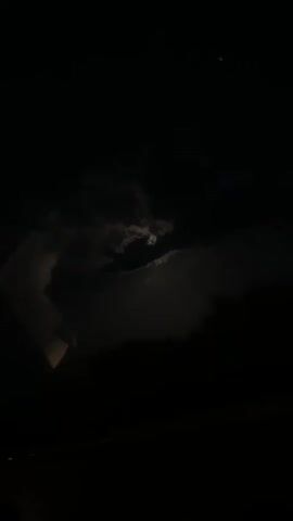 The video shows a dark night sky with the moon partially obscured by clouds. The moon is visible as a bright, glowing orb behind the clouds, creating an eerie and somewhat ominous atmosphere. There is a faint glow or illumination near the bottom left of the frame, possibly from a distant light source. The video appears to be shot from a slightly elevated position, as some rooftops or structures can be seen in the lower part of the frame. The overall impression is one of darkness, mystery, and a touch of the supernatural due to the obscured moon.