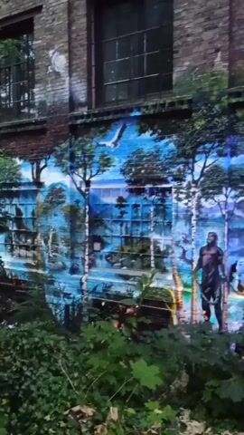 The video shows a mural painted on the side of a brick building. The mural depicts a fantastical landscape with a person standing in the foreground, a ship sailing on water in the background, and a creature resembling a lion with a woman on its back on the right side of the mural. There are trees and mountains in the background. The mural is vibrant and colorful, with blues, greens, yellows, and purples being the dominant colors. In the foreground, there is green foliage.