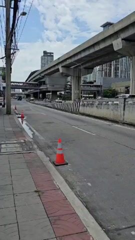 A POV video shows a street with a raised highway and buildings in the background. There are several cars and motorcycles driving on the road. The sky is cloudy. A traffic cone is visible on the sidewalk. The video shows buildings of varying heights, including high-rise apartments and commercial structures. The infrastructure appears to be urban and developed.