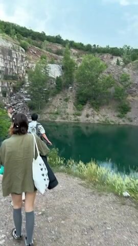 A group of young people, two females and one male, are walking along a dirt path next to a quarry with a deep blue-green lake at the bottom. The quarry walls are terraced and show signs of graffiti. The sky is partly cloudy. The group is moving from right to left in the initial shot, then the camera pans to show more of the quarry wall and the lake. One female is wearing a dark green top, a white tote bag with black heart designs, and gray knee-high socks. Another female has dark hair, a light-colored bikini top, and dark pants. The male has a white patterned shirt, dark pants, and a black sling bag. He is also carrying a rolled-up mat and a bottle.