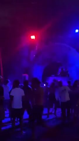 A crowd of people dances at an outdoor event at night. Blue and red spotlights illuminate the area. Behind the crowd is a large moon-shaped archway with a DJ booth inside. People are wearing casual clothing. The ground appears to be sandy.