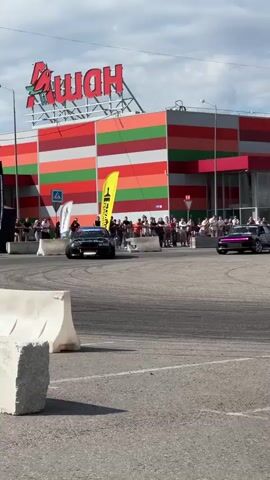 This video captures a dynamic drifting event taking place in a parking lot outside a large, modern retail building. The building, prominently featuring a large red sign with a bird logo and the Cyrillic word "Ашан" (Auchan), suggests a commercial setting. The facade of the building is a striking pattern of horizontal multicolored stripes in green, orange, and red. The sky is partly cloudy, indicating daytime.

The main subjects are two highly modified sports cars engaged in a drift competition or demonstration. Both cars are black and appear to be heavily customized for performance, with one adorned with intricate, colorful graphic art on its side. Tires are smoking as the cars execute controlled slides, kicking up dust and smoke, creating a visually exciting spectacle.

In the background, a crowd of spectators stands behind safety barriers, watching the event unfold. Several black and yellow flags, likely representing sponsors or teams, are visible in the area. White concrete barriers are strategically placed on the asphalt, marking the course or providing safety.

The overall atmosphere seems energetic and thrilling, as evidenced by the action of the cars and the presence of a watching audience. The location context of Vladimir, Russia, is supported by the visible Cyrillic text on the building and potentially on other signage which is less clear. The time of day is clearly daytime due to the bright lighting and shadows.