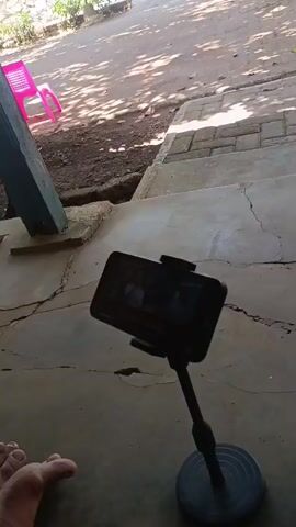 The video is taken from a low angle, showing a phone on a stand. The phone is playing a video or show, visible on the screen. Behind the phone, there are concrete steps leading up to a dirt area with a few pink chairs. The environment appears to be outdoors, with sunlight casting shadows on the ground. A person's foot is visible in the bottom left corner of the frame. The overall scene is rustic and casual.