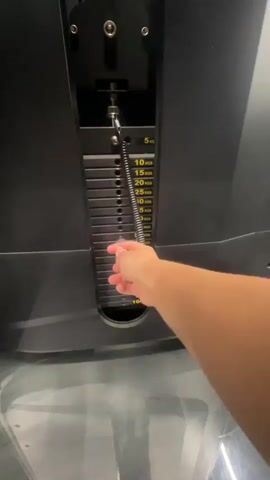 A close-up shot shows a person's hand adjusting the weight on a gym machine. The machine is black and has a stack of weights with yellow markings indicating kilograms. The hand inserts a metal pin into a slot to select a weight. The weights are labeled from 5 kg up to 100 kg. The scene appears to be indoors, likely a gym, with a gray floor visible. The lighting suggests it could be daytime, but no windows or natural light sources are apparent. The focus is entirely on the machine and the action of selecting the weight, with no clear indication of the person's emotions or the specific location beyond it being a gym. The time is indicated as 00:00-00:02 in the video.