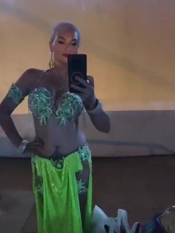 The video shows a woman with short, blonde hair and red lipstick posing in a mirror. She is wearing a bright green, sequined belly dancing costume consisting of a bra top, armbands, a hip belt, and a skirt with cutouts. The woman is holding a black smartphone in her right hand, angled to capture her reflection. The background appears to be indoors, with a warm-toned, possibly stuccoed wall and a reflective surface that may be a mirror or a glass table. There are some blurry objects in the foreground and background, including what looks like a metal railing on the left. The lighting is dim and casts a warm glow.
