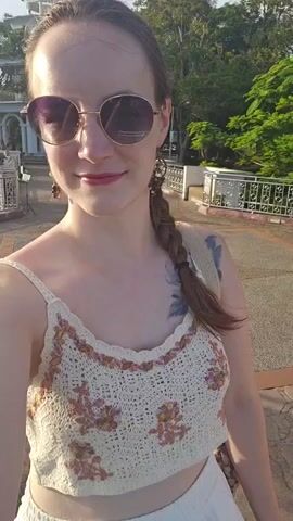 The video shows a young Caucasian woman with a braid in her hair and sunglasses. She is wearing a crocheted crop top and white shorts. She has a tattoo on her left shoulder. She is standing outside, on what appears to be a bridge or walkway. In the background, there are trees and buildings. Other people can be seen in the background. The sun is setting, casting a warm light on the scene.