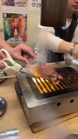 The video shows a person using scissors to cut a piece of meat being grilled over an open flame on a small, stainless steel grill. The meat is pink in the center, indicating it is rare. A second person is seated in the background. After being cut, the meat is placed on a metal plate next to a pair of chopsticks. A poster is visible on the wall behind the grill. The setting appears to be a restaurant.