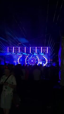 A nighttime outdoor concert or festival scene. A large stage is brightly lit with blue and white lights, featuring a central circular display with radiating patterns and vertical light bars. Laser lights, predominantly blue and some red, crisscross above the stage and the crowd. A dense crowd of silhouetted figures stands in front of the stage, some appearing to be dancing or milling about. In the foreground, a few individuals are more clearly visible, with one person on the left wearing a light-colored patterned shirt. The overall atmosphere is dark, illuminated by the stage and light show.