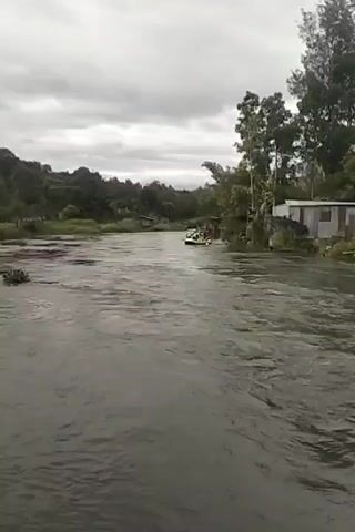 A wide shot shows a river with a jet ski on it. The river is flowing quickly. There are trees and houses on the banks of the river. The sky is cloudy.