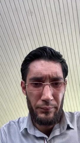 A man with dark hair and a beard wearing glasses and a collared shirt looks directly at the camera. The background consists of parallel, light-colored panels. The camera angle is from a low position, looking up at the man's face.