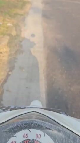 A first-person perspective video shows a road with a paved shoulder. To the left of the road is dry grass and some vegetation. To the right of the road is more vegetation and fields. In the distance, there are two people on a motorcycle. The sky is clear and blue.