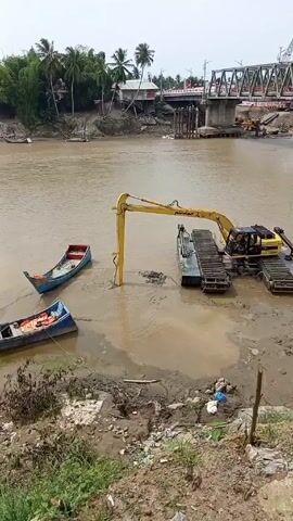 An excavator is working on a muddy river or lake bed on a grey day. The excavator is yellow with black accents, and is mounted on a floating platform. The arm of the excavator is extended into the water, and it appears to be dredging or moving mud. Two small boats are visible near the shoreline to the left of the excavator, one red and one blue, both appearing to be out of the water and resting on the shore. The water is murky and brown, suggesting the presence of sediment. In the background, a bridge spans the body of water, with visible construction or repair work underway. Palm trees and buildings are scattered along the opposite bank of the river, providing a sense of location. The camera is static, offering a steady view of the scene. The landscape is a mix of natural and man-made elements, highlighting ongoing construction activity in the area.