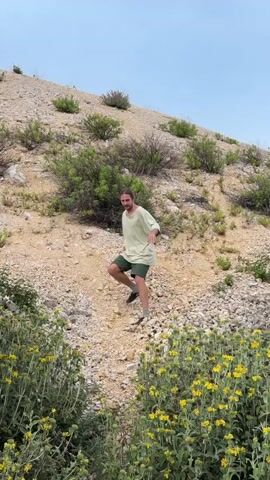 A man descends a rocky hillside covered with low-lying green vegetation and yellow flowers. He is wearing a light green shirt and dark green shorts, and has long hair. He walks with his arms outstretched for balance. The camera then pans upward to reveal a large wind turbine, its blades rotating in the sky behind the hill. The sky is overcast.