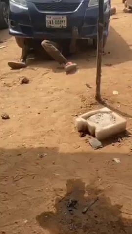 The video shows a man working on a blue Toyota car. The man is lying on the ground under the front of the car, apparently doing some kind of repair or maintenance. He is wearing brown pants and sandals. There is an oil stain on the ground near him, and a white object that looks like a container. The ground is dirt.  Another black car is parked nearby. He then gets up and stands in front of the car.