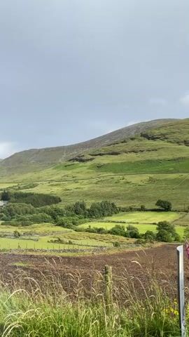 This video captures a scenic view of a lush green hillside under a cloudy sky. The foreground features a patch of tilled earth with tall grass and dry stalks, suggesting agricultural activity or a recently worked field. Beyond this, the landscape opens up to rolling green hills, dotted with trees and a few scattered buildings that appear to be houses. A stone wall delineates some of the fields, adding a rustic charm to the scene.

The sky is overcast with a muted, grey hue, but a faint rainbow arcs across the distant hills on the left side of the frame. This hints at recent rainfall or a passing shower. The overall impression is one of a tranquil, rural setting, likely in the United Kingdom given the location context. The lighting suggests it is daytime, perhaps late morning or afternoon, with the diffused sunlight from behind the clouds casting a soft glow on the landscape. There are no people visible in the frame, and the focus is entirely on the natural beauty of the countryside. The elements of the scene evoke a sense of peace and natural grandeur.