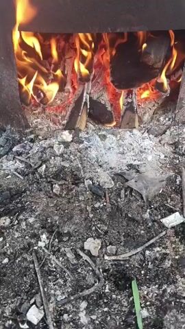 The video shows a close-up view of a fire burning inside a metal stove or brazier. Flames and glowing embers are visible, along with pieces of wood. The area in front of the stove is covered in ash, charcoal, and debris, including small sticks and what appears to be a piece of white paper or plastic. The lighting is dim, highlighting the brightness of the fire. The camera appears to be held steady, with minimal movement. There are no people visible in the frame, and no sounds are present. The overall impression is of an active fire in an outdoor or rustic setting.
