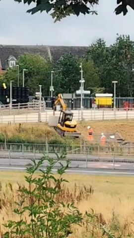 A yellow excavator is positioned on a dirt embankment, its arm extended upwards as if in operation. To the right of the excavator, two individuals in orange high-visibility vests and white hard hats are visible. The scene appears to be a construction or excavation site, with some fencing and debris in the background. The ground around the excavator is uneven and covered with dirt and vegetation. In the foreground, there is a patch of green foliage. A road is visible in the middle ground, with a barrier separating it from the construction area. The background shows buildings, trees, and what appears to be a railway station with platforms and overhead structures. The sky is overcast.