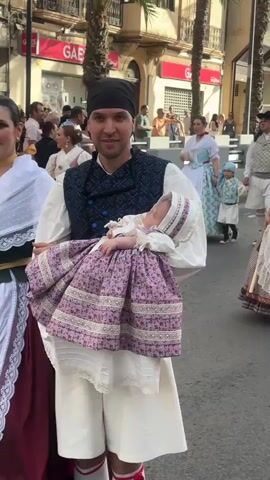 The video shows a street festival with people in traditional clothing. In the foreground, a man holds a baby dressed in a purple and white outfit. He is wearing a black headscarf, a dark blue patterned vest over a white shirt, and white knee-length pants with red garters. To his left, a woman stands smiling, wearing a white lace shawl over a white blouse, a dark red skirt, and a white apron with lace trim and a green sash. Behind them, other people in similar traditional attire are visible, as well as palm trees and buildings. The scene appears to be a procession or parade.