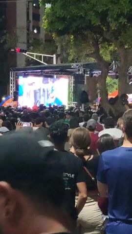 A medium shot shows a large crowd of people in front of an outdoor stage. The stage features a bright screen displaying video, and its supporting structure is decorated with white lights. The crowd is dense, with people packed close together. The majority of them face the stage, and some are holding their hands up in the air. Trees are visible behind the stage and to the sides. The people in the crowd are dressed in casual clothing and are diverse in appearance. The video appears to be shot from within the crowd, giving a sense of immersion in the event.