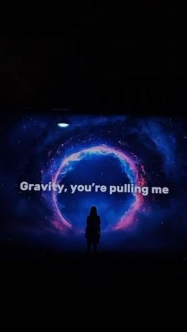 The video shows a television screen in a dark room. On the screen is an animated image of a person silhouetted against a background of a swirling galaxy. The lyrics "Gravity, you're pulling me through the stars and galaxies. Even when I fall apart" are displayed in white text on the screen.