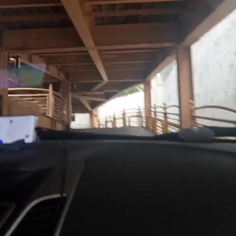 The video is filmed from the inside of a car, looking forward. The car is driving under a wooden structure with a sloped roof. A smartphone is mounted on the dashboard, displaying a navigation app. The exterior of the car's interior, including the dashboard, steering wheel, and air vents, are visible. The environment appears to be outdoors, with greenery visible in the background. The video is short, lasting only a few seconds.