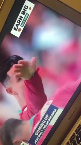 A person is shown on a laptop screen. They are wearing a pink jersey with a logo. There are two superimposed ads on the screen. One ad is in the top left corner and says "EARN BIG CLICK HERE". The other ad is in the bottom right corner and says "COMING UP JUST WHAT THE DR ORDERED". A QR code is also visible. The person appears to be a male athlete, possibly a soccer player, with dark hair and a shaved head. Their facial expression is one of frustration or exhaustion. The background is blurred and appears to be a crowd or stadium. The laptop is open and the keyboard is partially visible. The video is shaky and tilted.