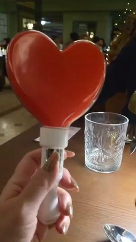 A hand with gold glitter fingernails holds a heart-shaped, light-up wand on a table with a small, clear glass tumbler and silver cutlery. The wand is red and emitting light from its center. The table is made of wood. There are blurred figures in the background, including some Christmas lights. The wand is switched off and then on again.