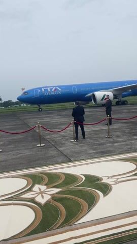 The video shows a blue ITA Airways airplane parked on a tarmac.  In front of the plane, there is a red velvet rope barrier held up by gold stanchions. Two men in suits stand behind the barrier, facing the plane. One of the men is holding a camera, and it appears he is photographing the plane. In the foreground, a patterned rug can be seen. The sky is overcast and the overall lighting is somewhat muted.