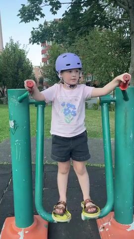 A young child wearing a purple helmet, a pink t-shirt with a blue floral design, and dark shorts is standing on a piece of outdoor exercise equipment in a park. The child has their hands on the red handles of the equipment and their feet on the green pedals, which are attached to orange base supports. The child is looking forward and appears to be moving their legs back and forth on the pedals. The background shows green grass, trees, and a red apartment building.