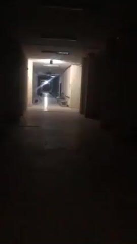 The video begins in a dimly lit, narrow hallway. The camera is positioned at one end, looking towards a brighter area at the opposite end. The walls of the hallway are a light color. Towards the bright end, some objects, possibly construction materials or furniture, are visible. After approximately three seconds, the camera abruptly turns to the right, plunging the view into darkness. The video ends with only blackness visible.