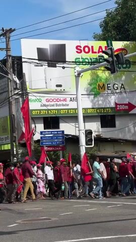 A group of people are walking across a street in a daytime shot. Most are carrying red flags. The majority appear to be men, although some women are in the group as well. Most of the people in the group are wearing red hats. They are walking from left to right in the frame. A building painted yellow is visible behind them with various flags attached to it. Traffic lights are visible above and to the side of the people. A sign for "Mega Solar" is visible behind the traffic light.