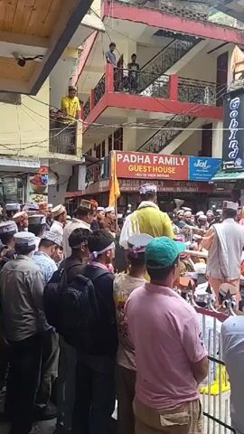 This video captures a vibrant daytime street scene in Kasol, India, featuring a procession or religious gathering. Numerous people, predominantly men, are present, with many wearing traditional Himachali caps. A central object, possibly a deity or palanquin, is elaborately decorated with yellow, purple, and white fabrics and garlands. This object is surrounded by a dense crowd on the street level.

Some individuals are visible on the balconies of nearby buildings, observing the event from above. A large metal horn or trumpet can be seen among the crowd, suggesting that music is part of the procession. The atmosphere appears to be one of observance and engagement.

The setting is a narrow street lined with multi-story buildings that house commercial establishments. Visible signs include:
- "PADHA FAMILY GUEST HOUSE - RIVER VIEW ROOMS"
- "HOT BATH WITH RIVER VIEW ROOMS RESTAURANT"
- "Jal - Joy of India - Bath Fittings & Bathroomware"
- "Gobind Garments & Shawls - 100% PURE"

The weather appears clear and bright, indicating a sunny day.