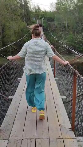 A woman with her hair in a bun walks away from the camera on a wooden suspension bridge. She wears a light striped shirt, teal pants and yellow sandals. Her arms are outstretched for balance.  The bridge has wire mesh sides. Other people are visible in front of her on the bridge. Trees surround the bridge, and the sky is cloudy. The wind appears strong, moving the trees.