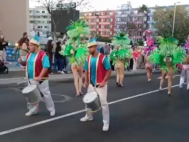 The video shows a carnival parade taking place on a city street. Two men are in the foreground, both wearing hats, red vests, blue shirts, and white pants. They are each playing a drum and walking in the same direction. Behind them is a group of women in colorful carnival costumes, primarily green and pink, featuring elaborate feathered headdresses. Some of the women are wearing minimal clothing and their backs are exposed. There is a crowd of people on the sidewalks watching the parade, including families with strollers. The buildings behind the crowd are multi-story and painted in various colors. The video appears to be taken from a low angle, emphasizing the movement and energy of the parade.