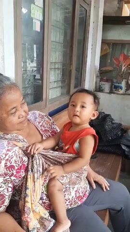 A medium shot shows an elderly woman and a baby sitting on a wooden bench in front of a window. The woman is on the left and the baby is on her lap. The woman has gray hair and is wearing a colorful floral print shirt. The baby is facing the camera and is wearing an orange tank top and a patterned cloth around its waist and legs. The baby is looking to the right. The woman is looking down at the baby. The background consists of a window with wooden frames, a wall, and some plants. The lighting is natural and appears to be daylight.