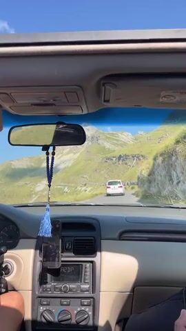 The video shows a first-person perspective from inside a car driving on a mountain road. The camera is looking forward through the windshield and at the dashboard. The road is winding and ascends a grassy, rocky mountainside. Another white car is visible ahead on the road. The sky is clear and blue. The interior of the car is visible, including the steering wheel, dashboard, rearview mirror with a dangling blue ornament, and a mobile phone mounted on the dashboard. A person's arm is visible on the left side of the frame, holding the steering wheel. Another person's head and shoulder are visible in the lower right corner of the frame, holding a phone. The camera also briefly pans to the right, showing the side window and the mountainous terrain outside. The overall scene is a scenic drive through mountains on a sunny day.